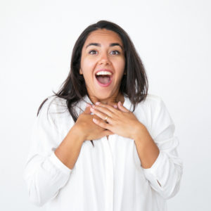 Happy shocked woman gasping of surprise. Joyful beautiful young Latin woman in white shirt keeping hands on chest, smiling and shouting for joy. Good news and surprise concept